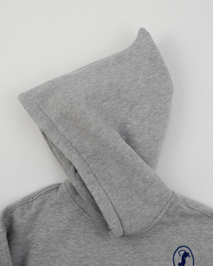 A close-up of a heather gray hoodie with the hood pulled up, featuring a small navy blue circular logo on the left chest, shot against a plain light gray background.