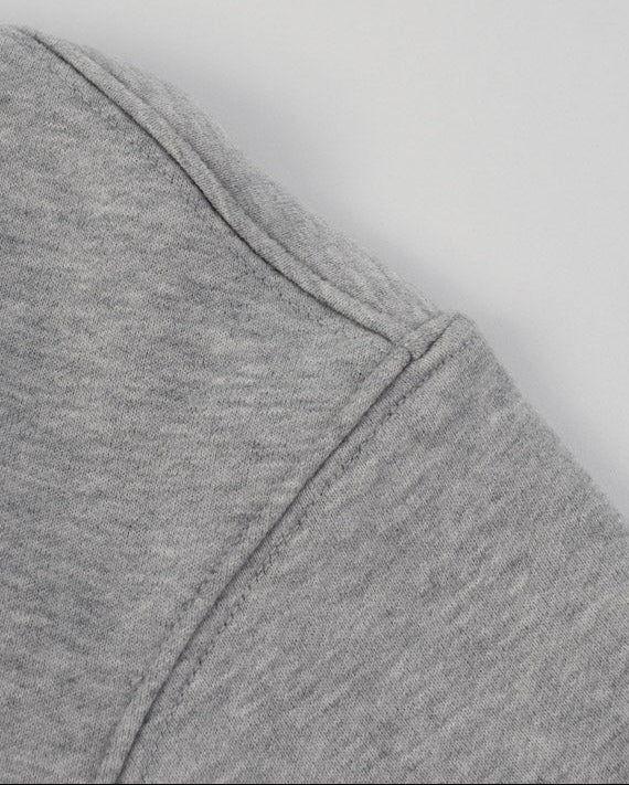 Close-up of a heather gray sweatshirt showing the shoulder seam and a navy blue embroidered circular logo on the upper left chest, set against a plain light gray background.