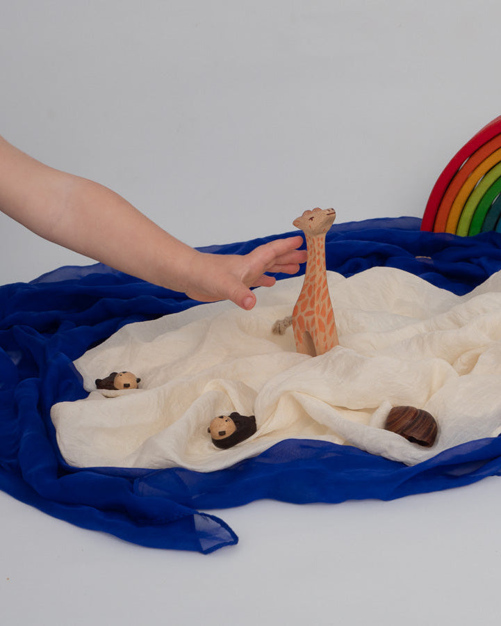 A wooden giraffe toy is held by a person's hand over a styled play scene with a cream fabric island, blue fabric ocean, small wooden animals, and a rainbow arch toy on a white background.